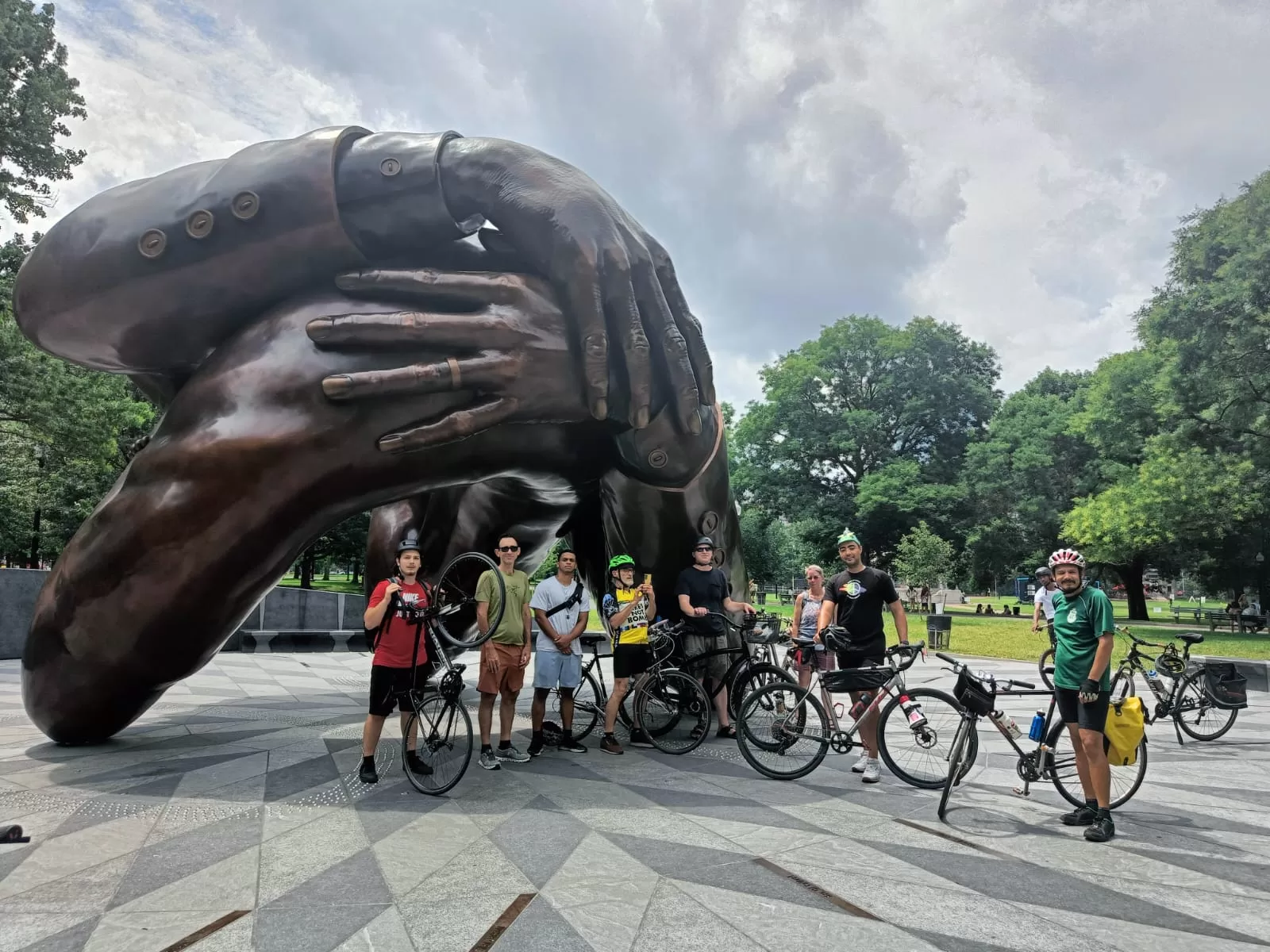 A small group of cyclists stand in front of the "Embrace" sculpture in Boston Common.