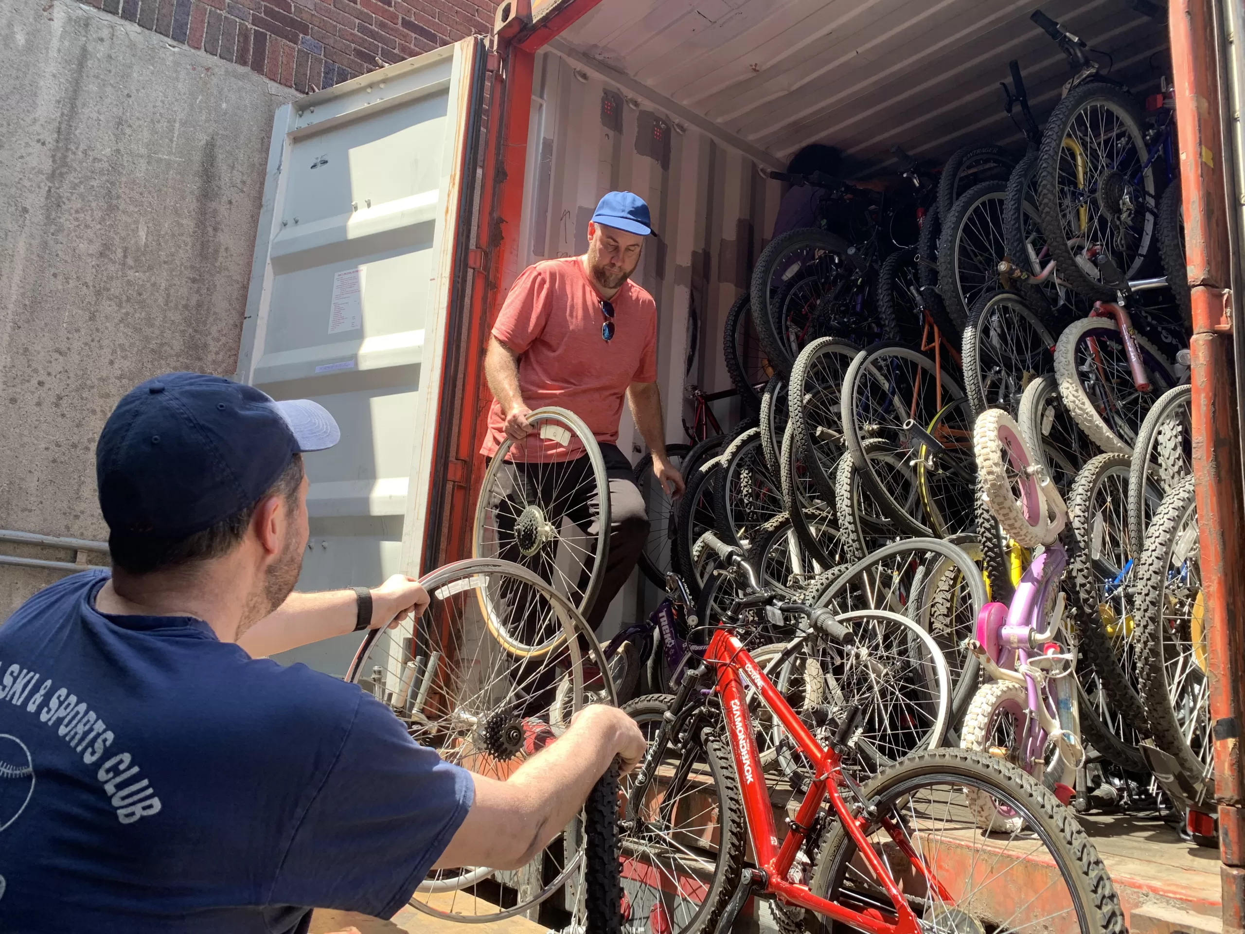 A man hands a bike to a second man, who is standing at the opening of a shipping container with hundreds of bikes inside of it.