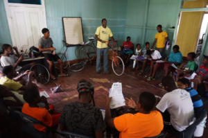 A group of young adults and youth watch an instructor during an Earn-A-Bike lesson