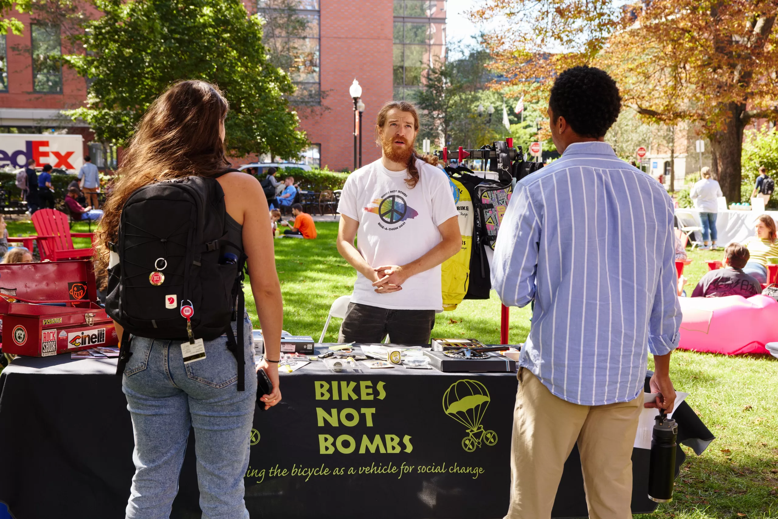 A white man stands behind a booth labeled "Bikes Not Bombs" and talks to a couple of college students.